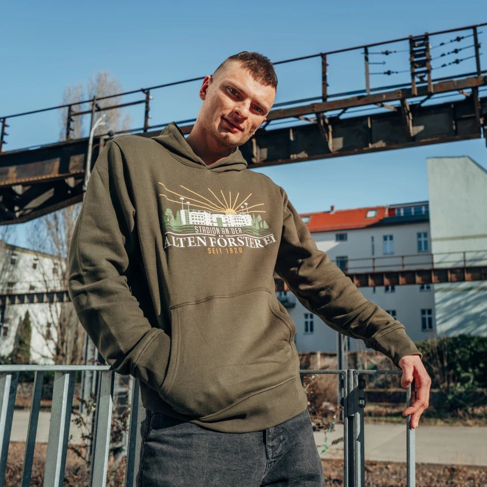 Junger Mann in grüner Kapuzenjacke steht vor einer Metallkonstruktion und blickt in die Kamera. Urbanes Umfeld.