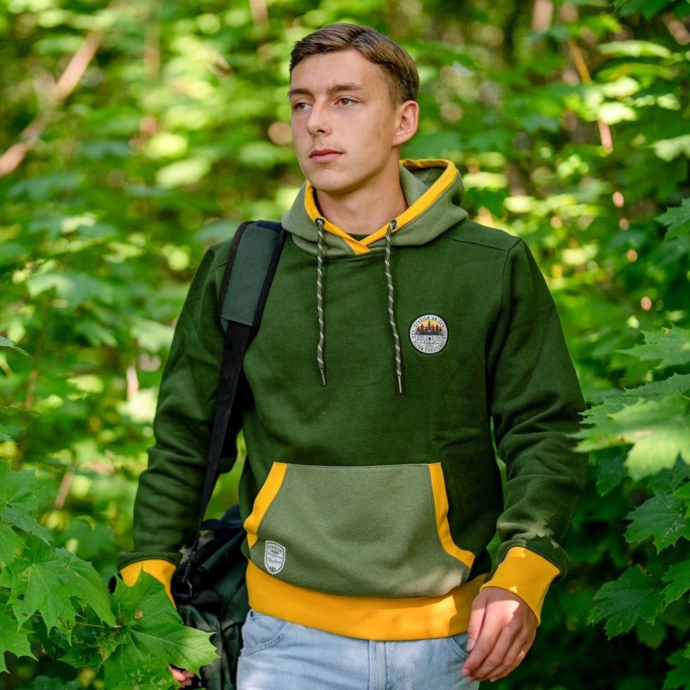 Junger Mann in grüner Jacke mit gelben Akzenten steht in einem grünen Wald. Er trägt eine Tasche über der Schulter.
